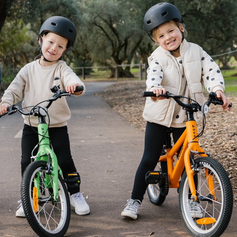 Two young children wearing black helmets ride bicycle bikes on a paved path in a park. The child on the left rides a green bike with a beige sweatshirt and black pants, while the child on the right rides an orange bike with a beige puffer vest with black polka dots, black pants, and sneakers.