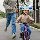 A man in a hoodie and blue jeans runs alongside a young girl wearing a pink helmet and riding a purple children's bicycle on an outdoor paved path.