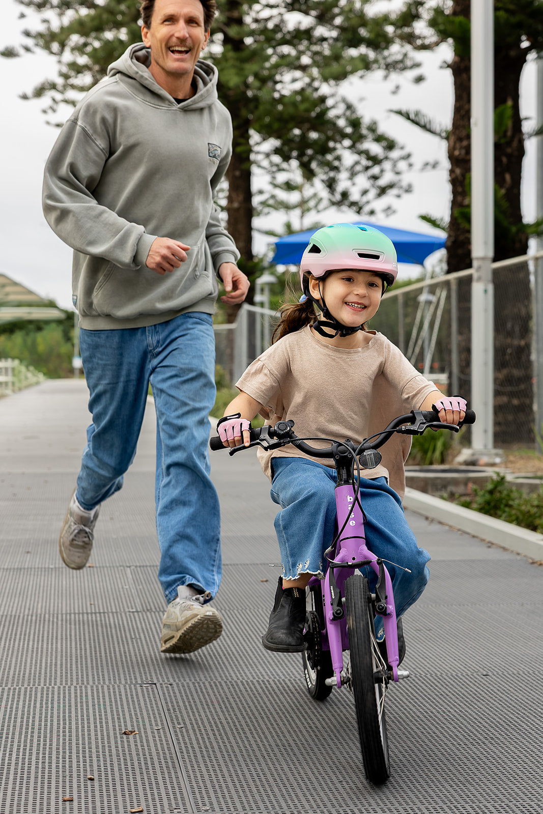 A man in a hoodie and blue jeans runs alongside a young girl wearing a pink helmet and riding a purple children's bicycle on an outdoor paved path.