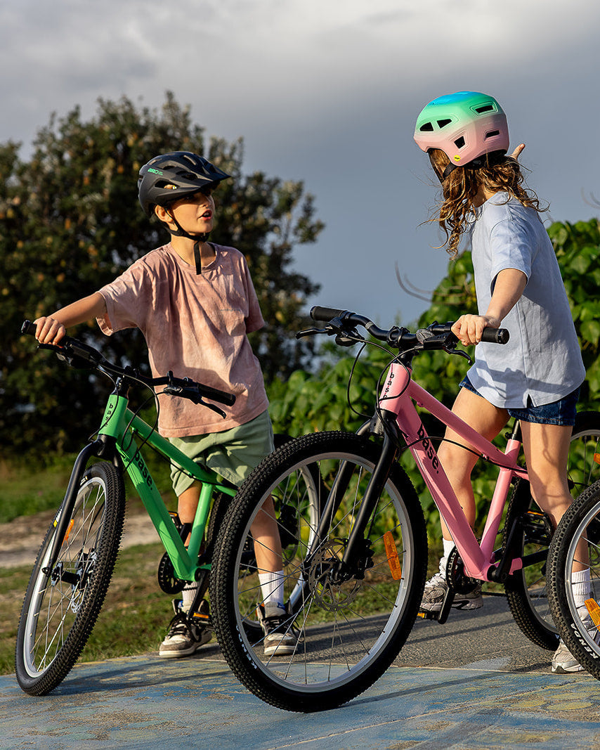 Three children wearing helmets stand with their bikes outdoors on a paved path with green trees in the background. The bikes are green and pink.