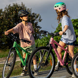 Three children wearing helmets stand with their bikes outdoors on a paved path with green trees in the background. The bikes are green and pink.