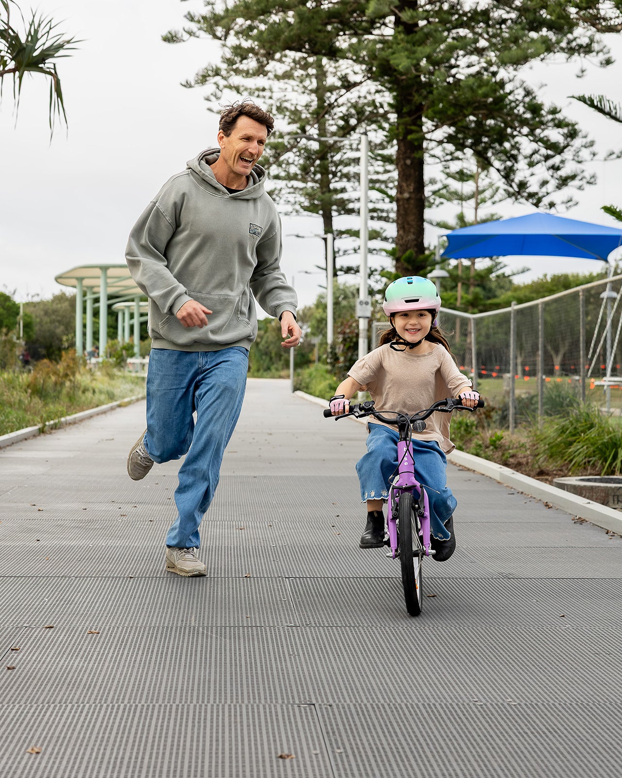 A man in a hoodie and blue jeans runs alongside a young child on a purple bike wearing a mint green helmet, on an outdoor paved path.