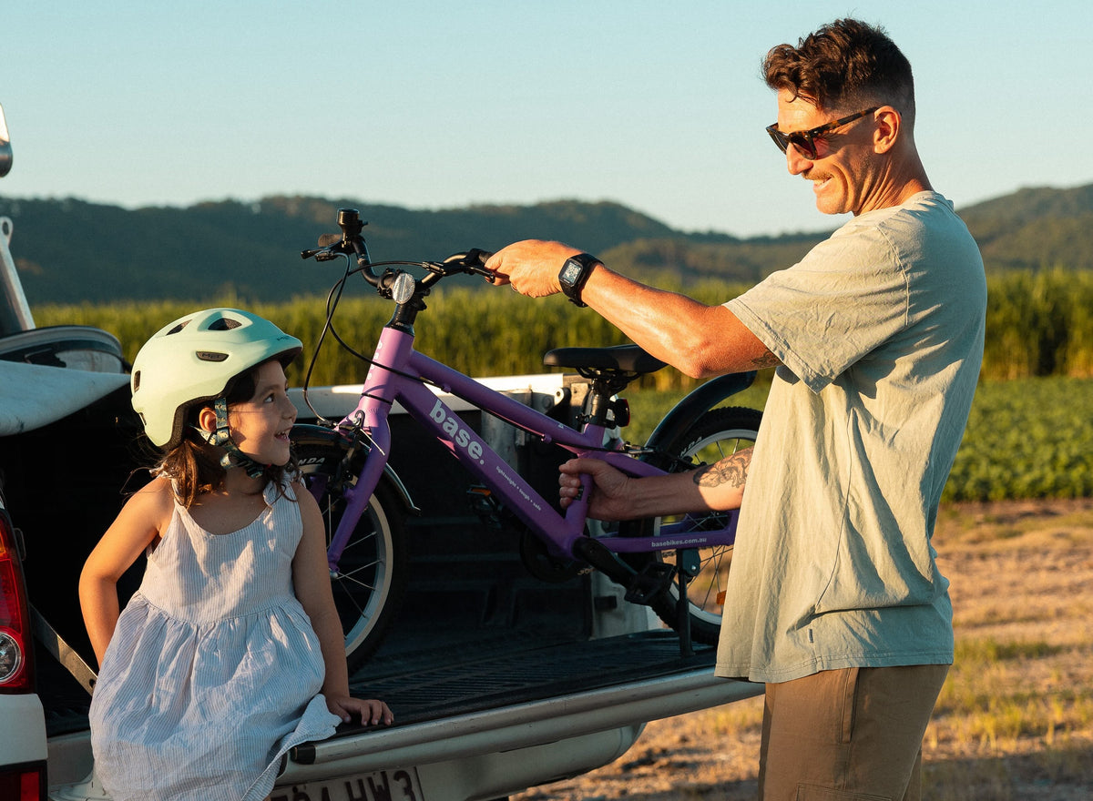 A man and a young girl unload a purple mountain bike from the bed of a ute outdoors. The man wears sunglasses and a light grey t-shirt, and the girl wears a white dress and a light green bicycle helmet.