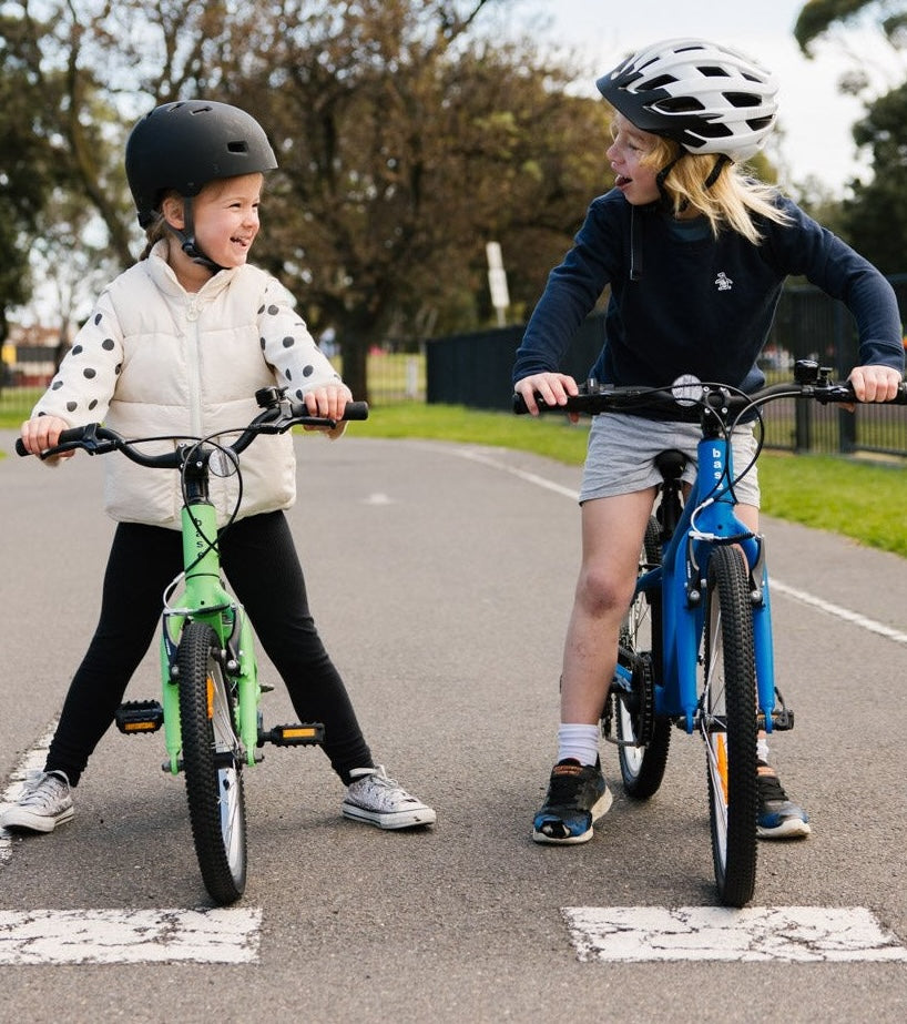 Two children riding bicycles on a paved path with trees in the background