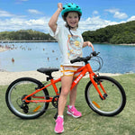 A young girl wearing a green helmet and a white graphic t-shirt stands next to an orange 20 inch bike on a grassy lawn in front of a beach and blue water.