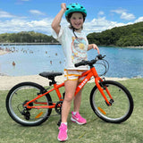 A young girl wearing a green helmet and a white graphic t-shirt stands next to an orange 20 inch bike on a grassy lawn in front of a beach and blue water.