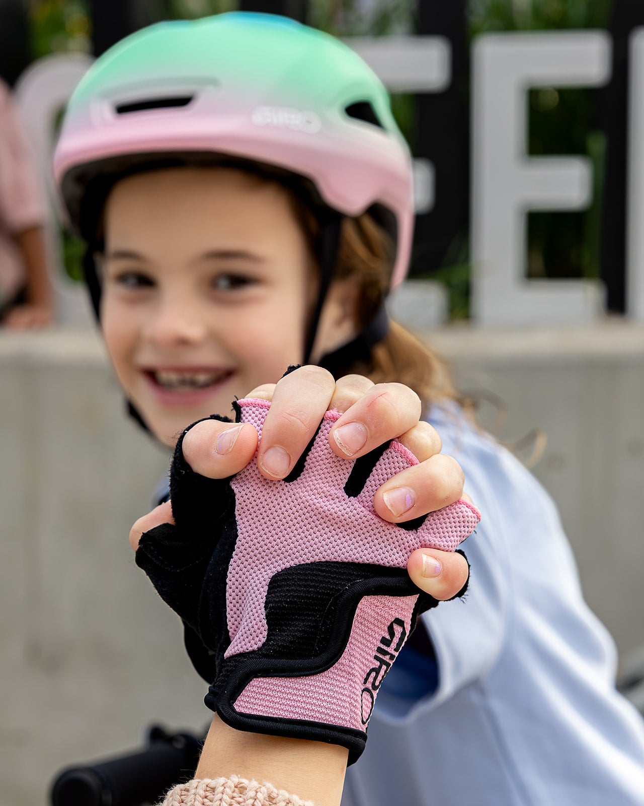 A child wearing a Giro helmet and pink and black fingerless cycling gloves, with the child's hands prominently featured in the foreground against a blurred outdoor background.
