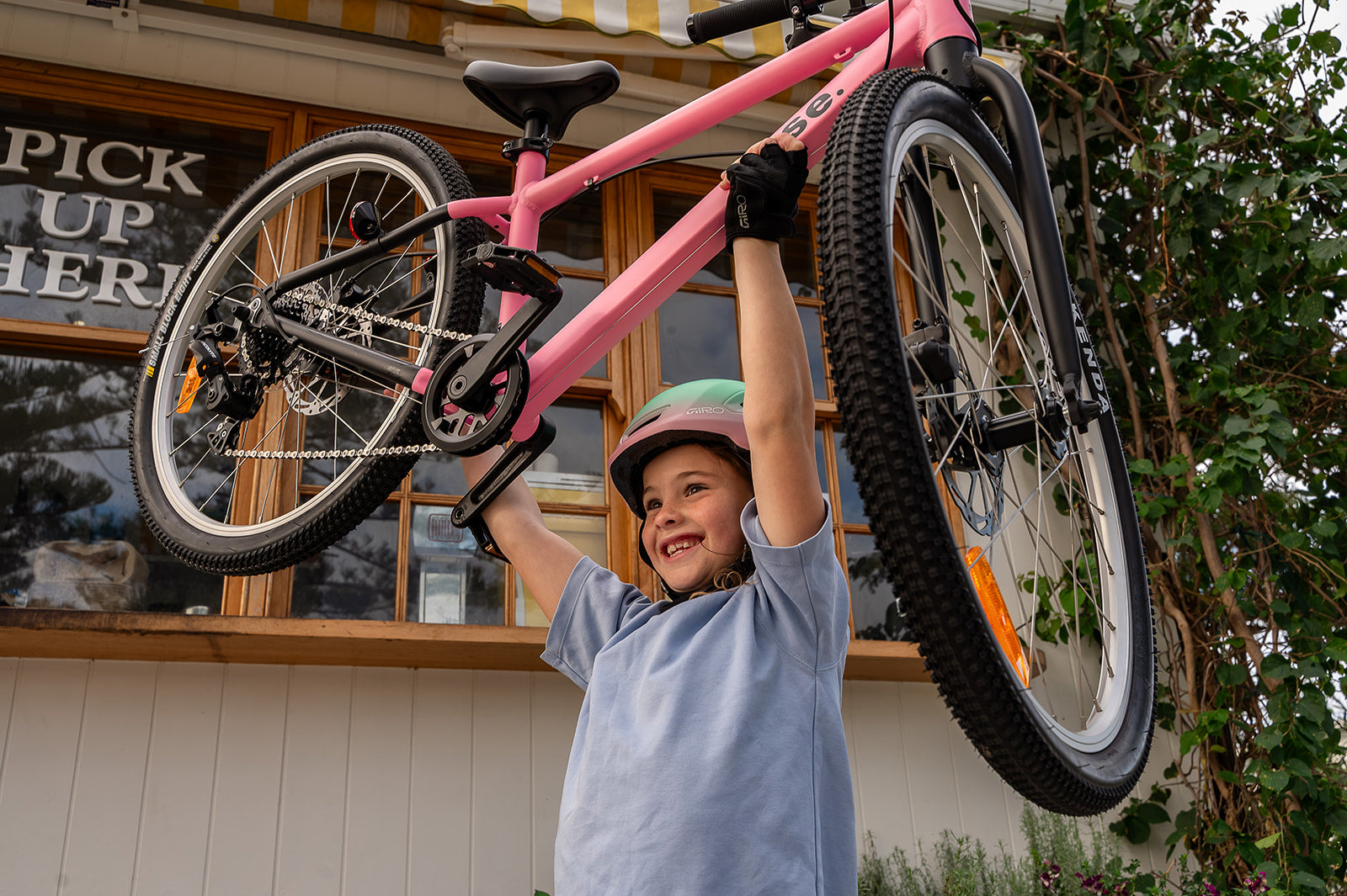 A young girl wearing a green helmet smiles as she lifts a pink children's bicycle above her head outside a shop.
