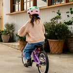 A child wearing a pink and mint green bicycle helmet, a beige knit sweater, and blue jeans rides a purple children's bicycle on a concrete path in front of a white house with large windows and potted plants.