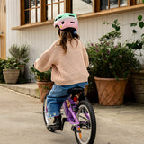 A child wearing a pink and mint green bicycle helmet, a beige knit sweater, and blue jeans rides a purple children's bicycle on a concrete path in front of a white house with large windows and potted plants.