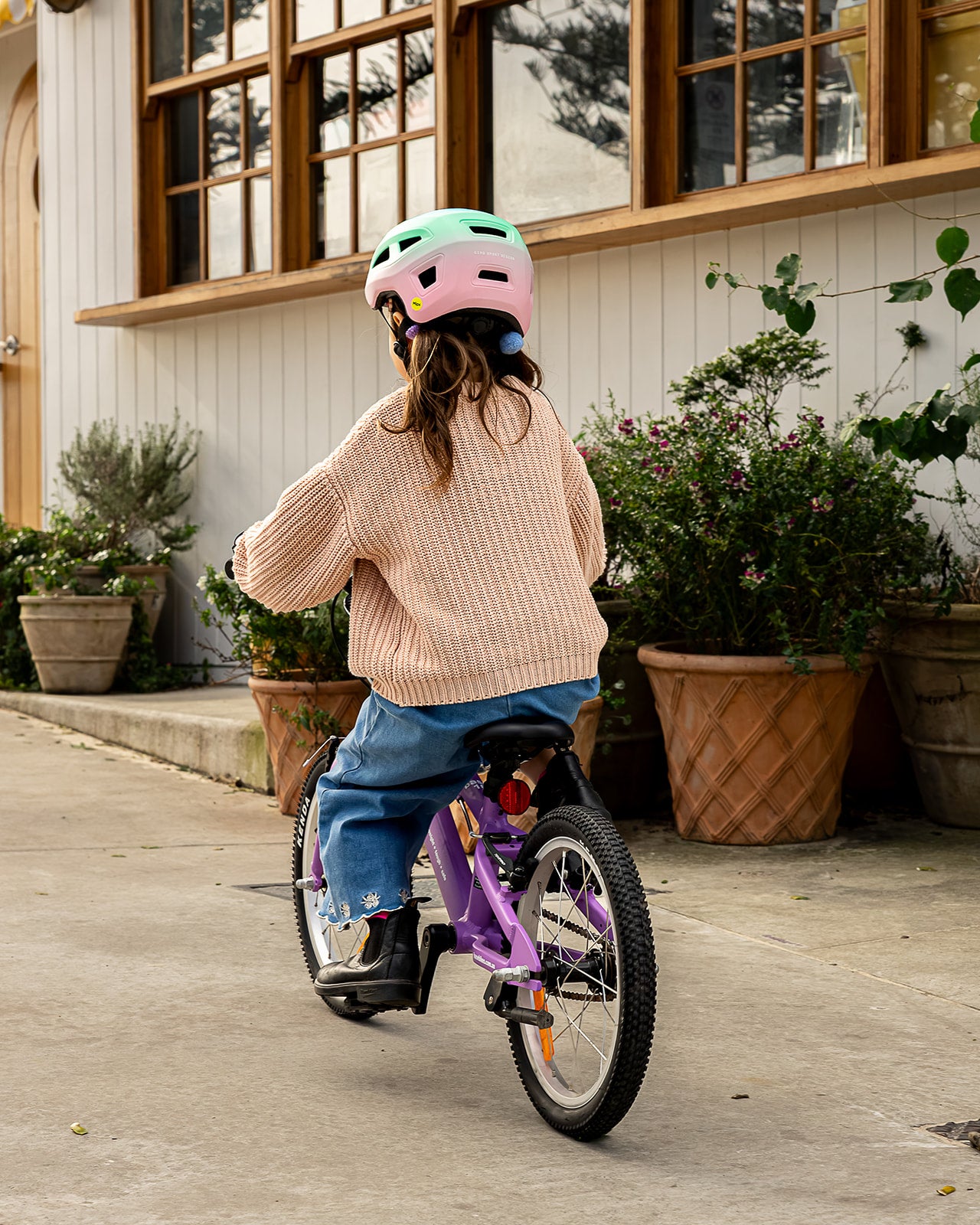 A child wearing a pink and mint green bicycle helmet, a beige knit sweater, and blue jeans rides a purple children's bicycle on a concrete path in front of a white house with large windows and potted plants.