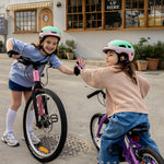 Two young girls wearing helmets and gloves high-five while riding small bicycles in front of a coffee shop. One bike is pink and the other is purple, set on a paved street with storefront windows in the background.