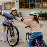 Two young girls wearing helmets and gloves high-five while riding small bicycles in front of a coffee shop. One bike is pink and the other is purple, set on a paved street with storefront windows in the background.