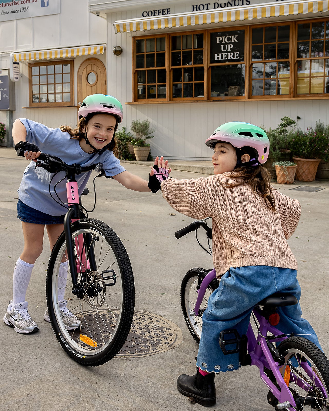 Two young girls wearing helmets and gloves high-five while riding small bicycles in front of a coffee shop. One bike is pink and the other is purple, set on a paved street with storefront windows in the background.