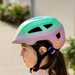 A child wearing a Giro bicycle helmet with a pastel green, pink, and purple gradient. The helmet features black vents, a black chin strap, and a small yellow MIPS safety sticker.