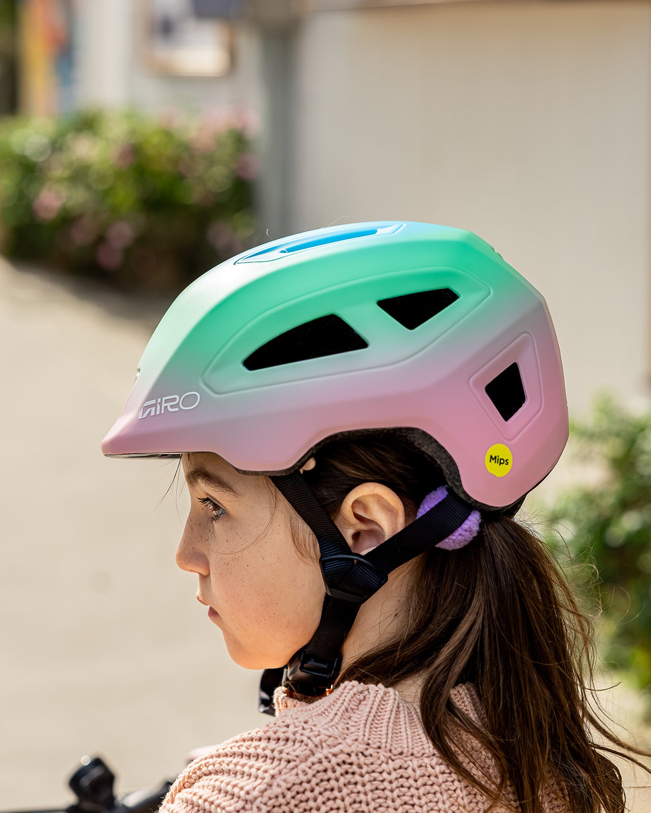 A child wearing a Giro bicycle helmet with a pastel green, pink, and purple gradient. The helmet features black vents, a black chin strap, and a small yellow MIPS safety sticker.