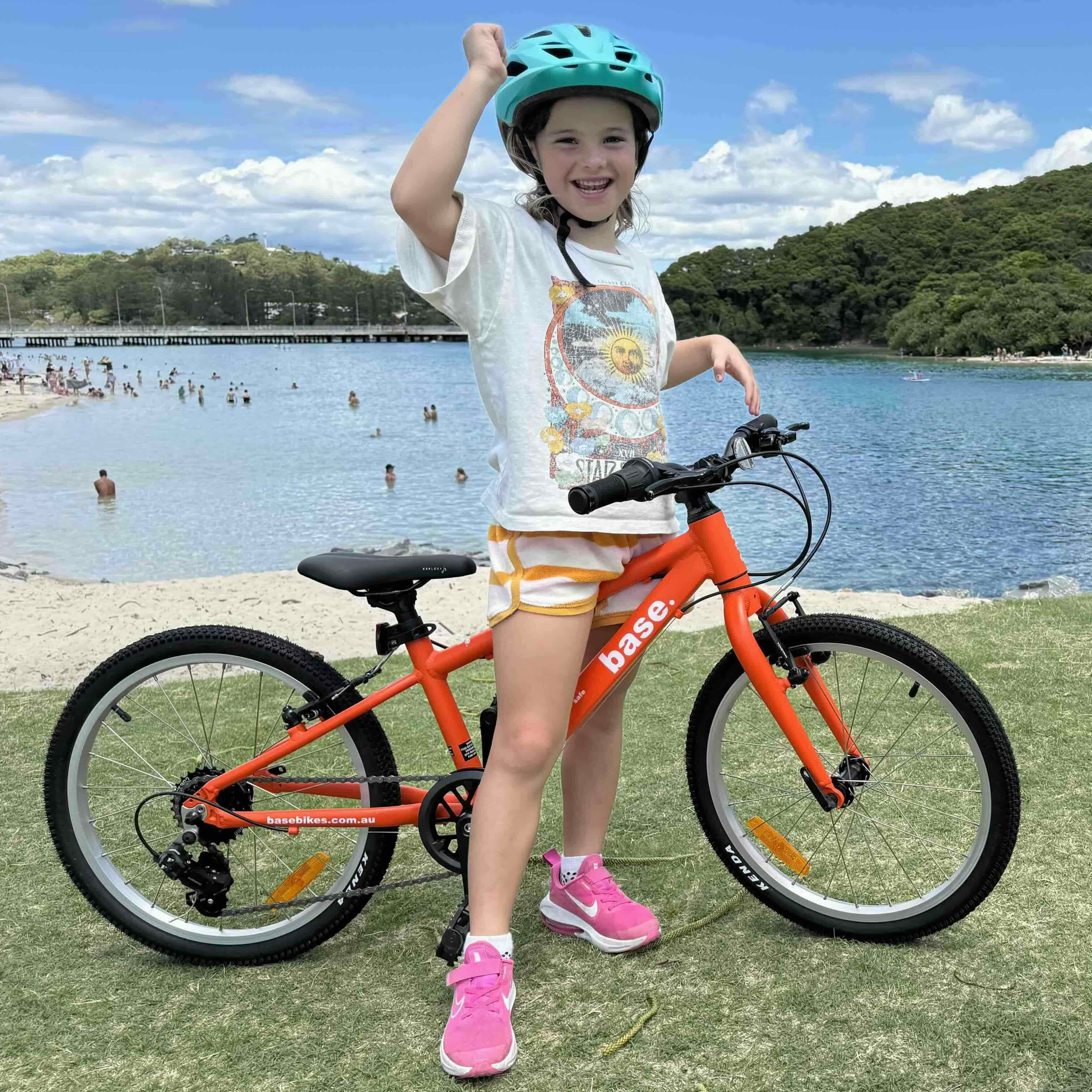A young girl wearing a green helmet and a white graphic t-shirt stands next to an orange base kids' bicycle on a grassy lawn in front of a beach and blue water.