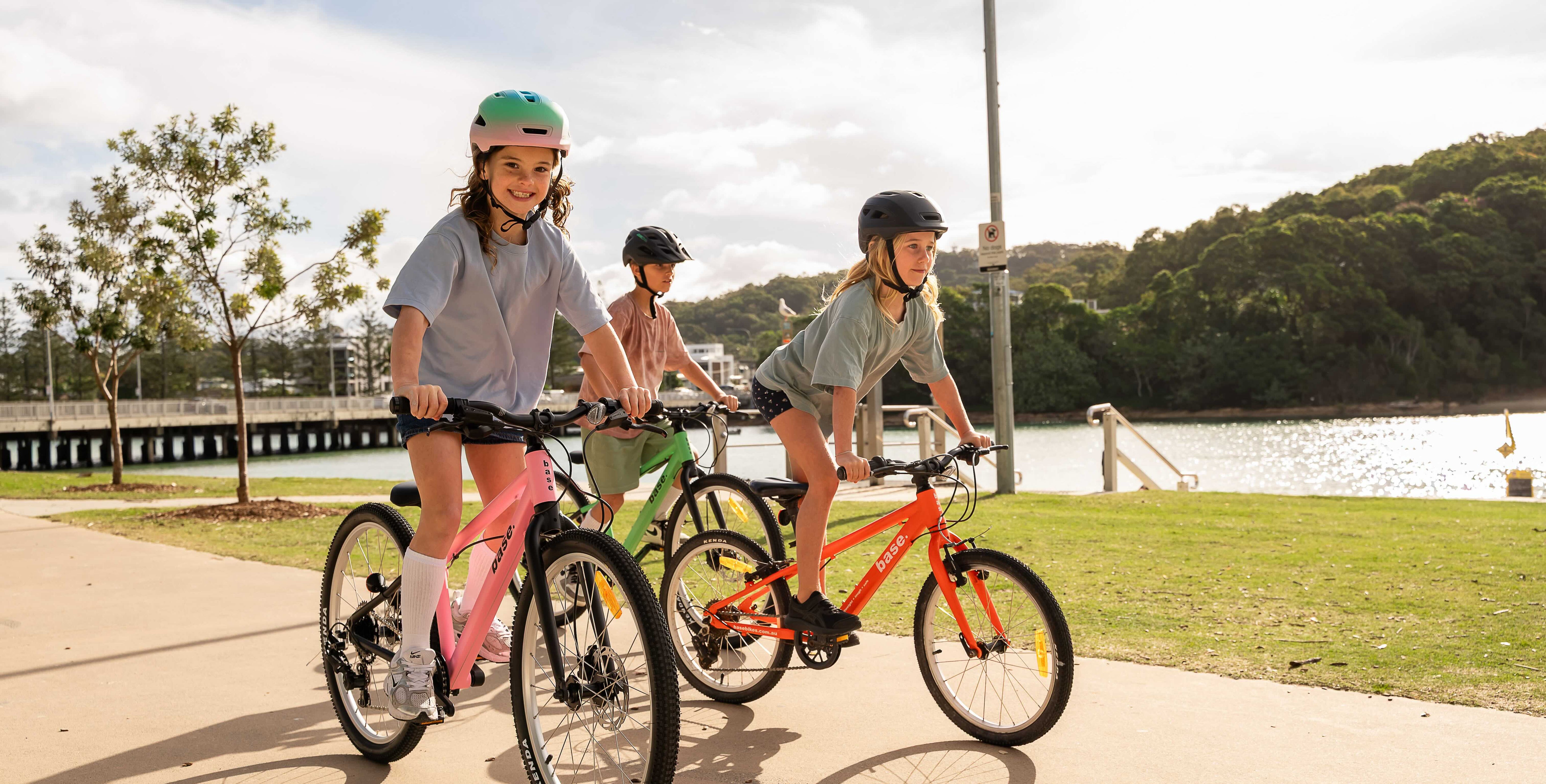 Three children riding colorful bicycles on a paved path next to a lake with trees in the background.