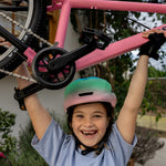 A smiling child wearing a pink and green giro bicycle helmet and a light blue t-shirt holds up a pink bicycle outdoors. The child is wearing black giro fingerless gloves.