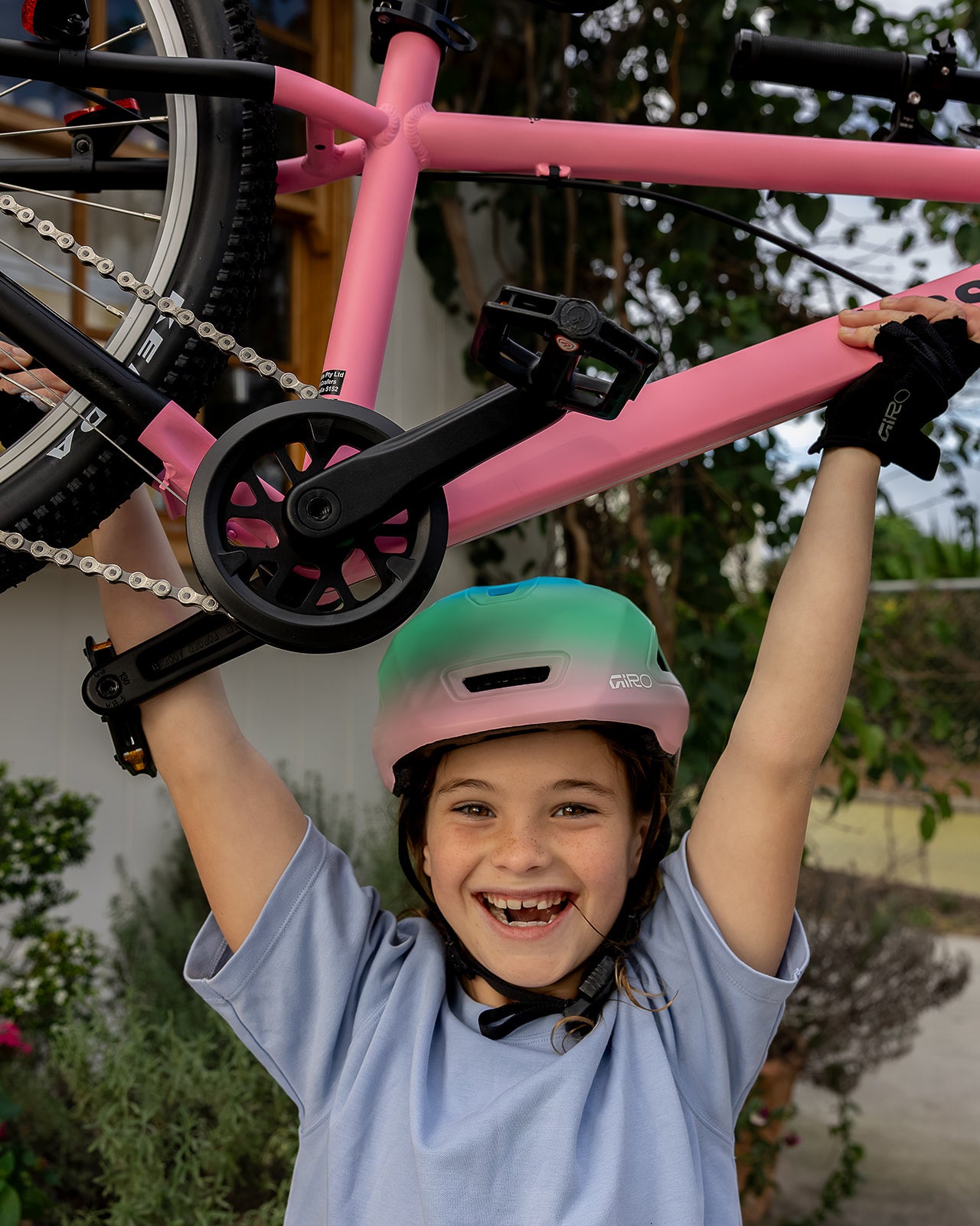 A smiling child wearing a pink and green giro bicycle helmet and a light blue t-shirt holds up a pink bicycle outdoors. The child is wearing black giro fingerless gloves.