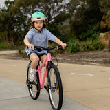 A young girl wearing a mint green and pink bicycle helmet rides a pink children's bicycle on an outdoor paved path with trees in the background.