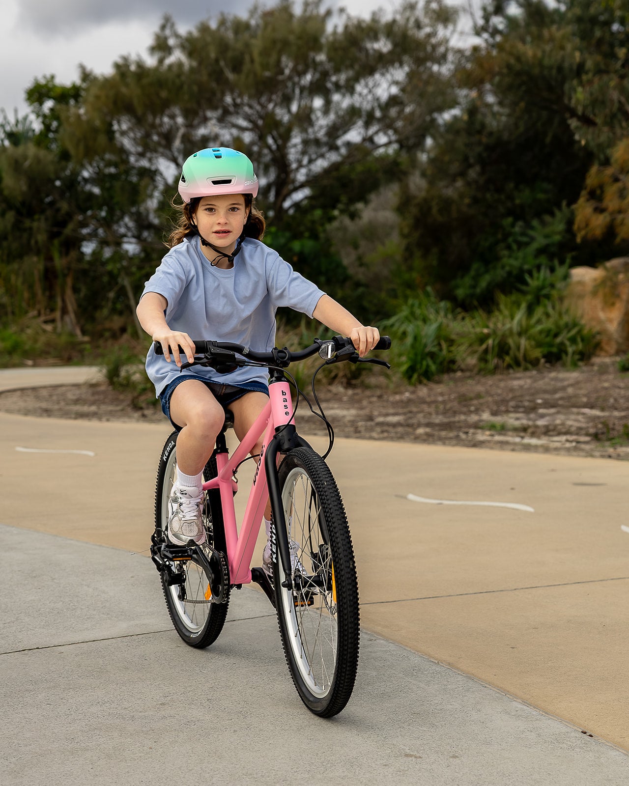 A young girl wearing a mint green and pink bicycle helmet rides a pink children's bicycle on an outdoor paved path with trees in the background.