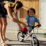 A woman assists a young boy learning to ride a white base balance bike outdoors in front of a wooden door.