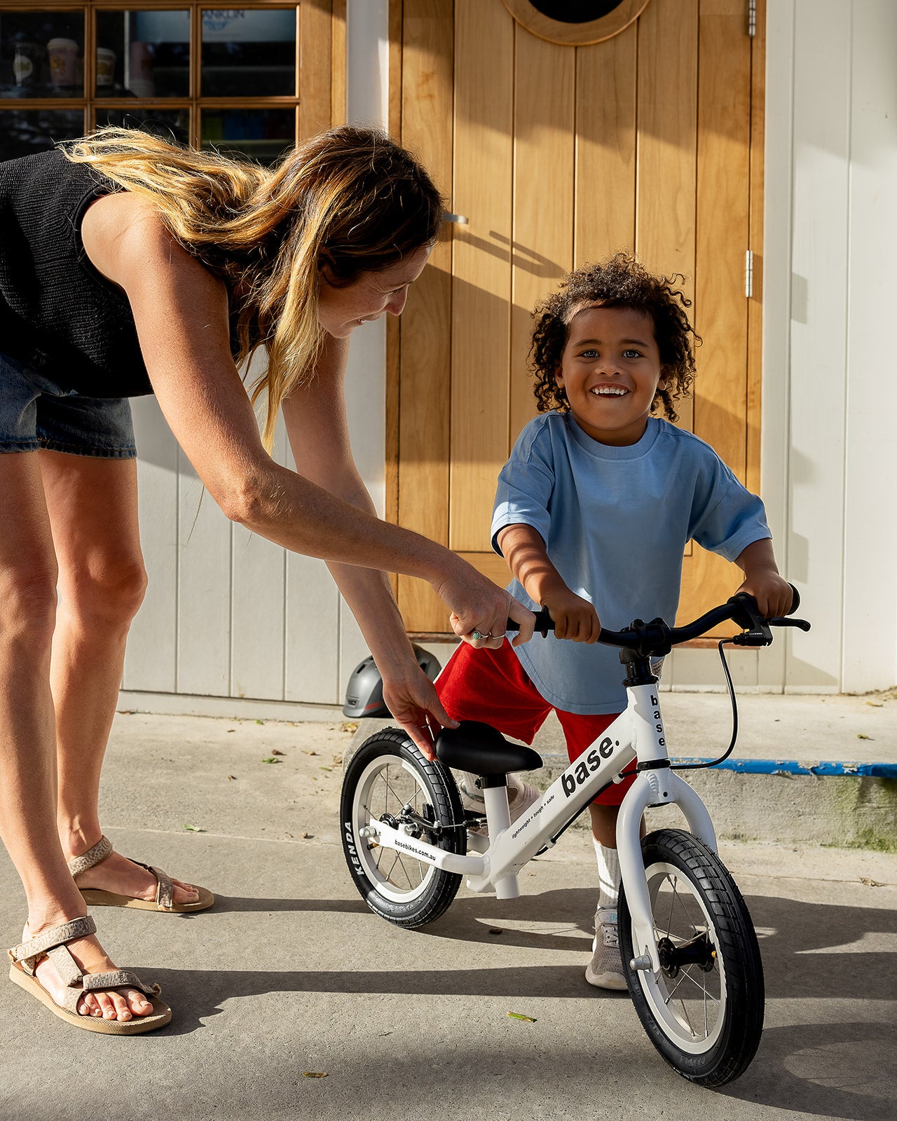 A woman assists a young boy learning to ride a white base balance bike outdoors in front of a wooden door.