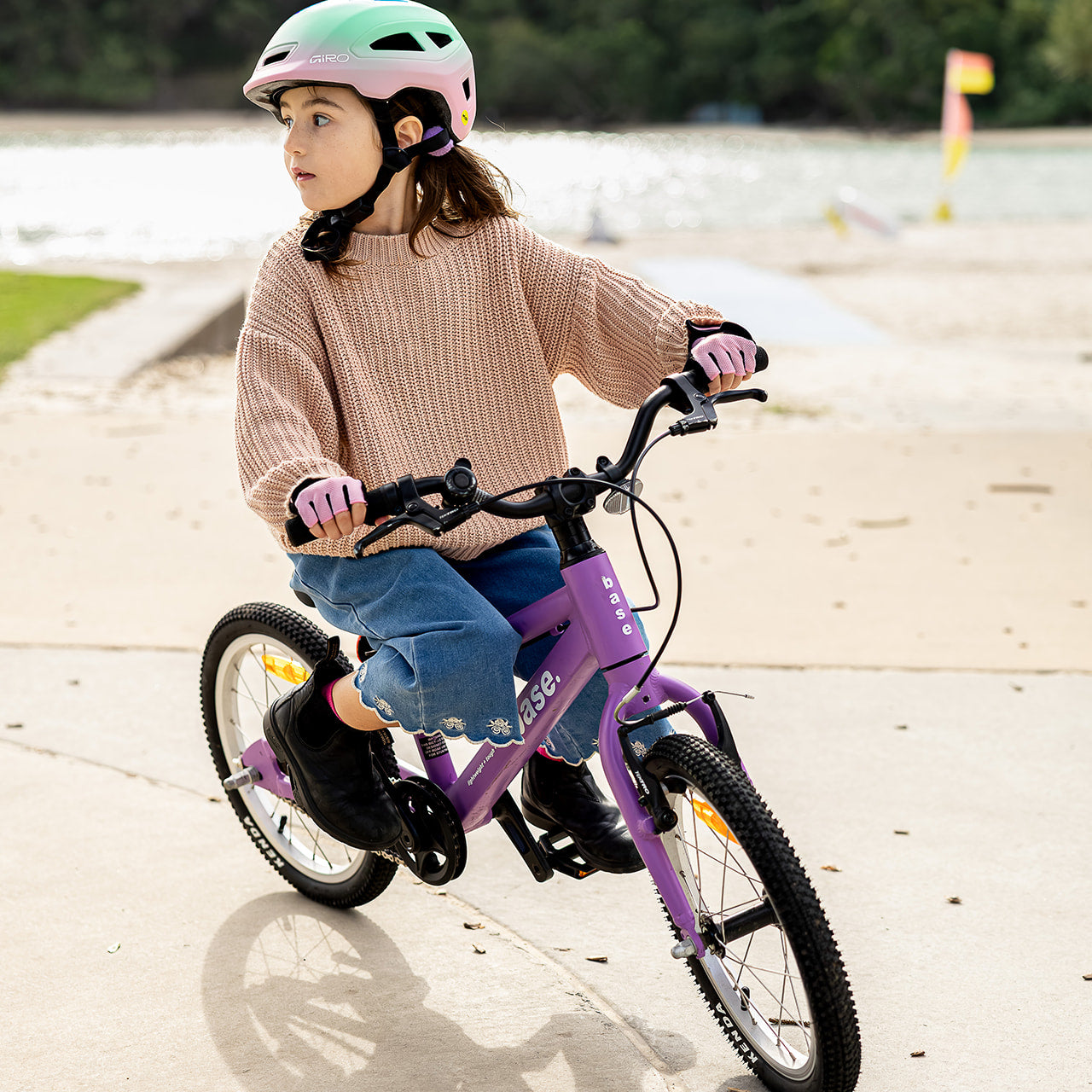 A young child wearing a mint green and pink helmet rides a purple kids' bicycle on a concrete path near a beach or lake, with trees in the background.