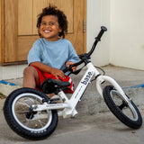 A smiling young child sits by a white balance bike.
