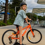 A young girl wearing a black helmet and a light blue t-shirt stands next to an orange children's bicycle in an outdoor paved area with trees and a shelter in the background.