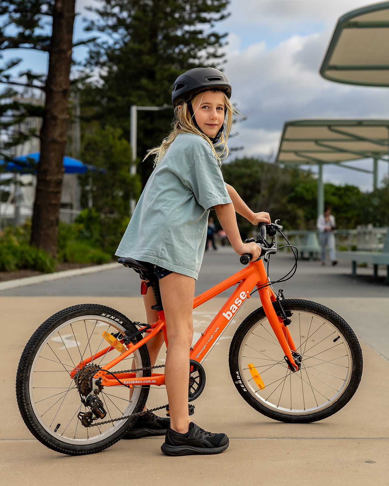 A young girl wearing a black helmet and a light blue t-shirt stands next to an orange children's bicycle in an outdoor paved area with trees and a shelter in the background.