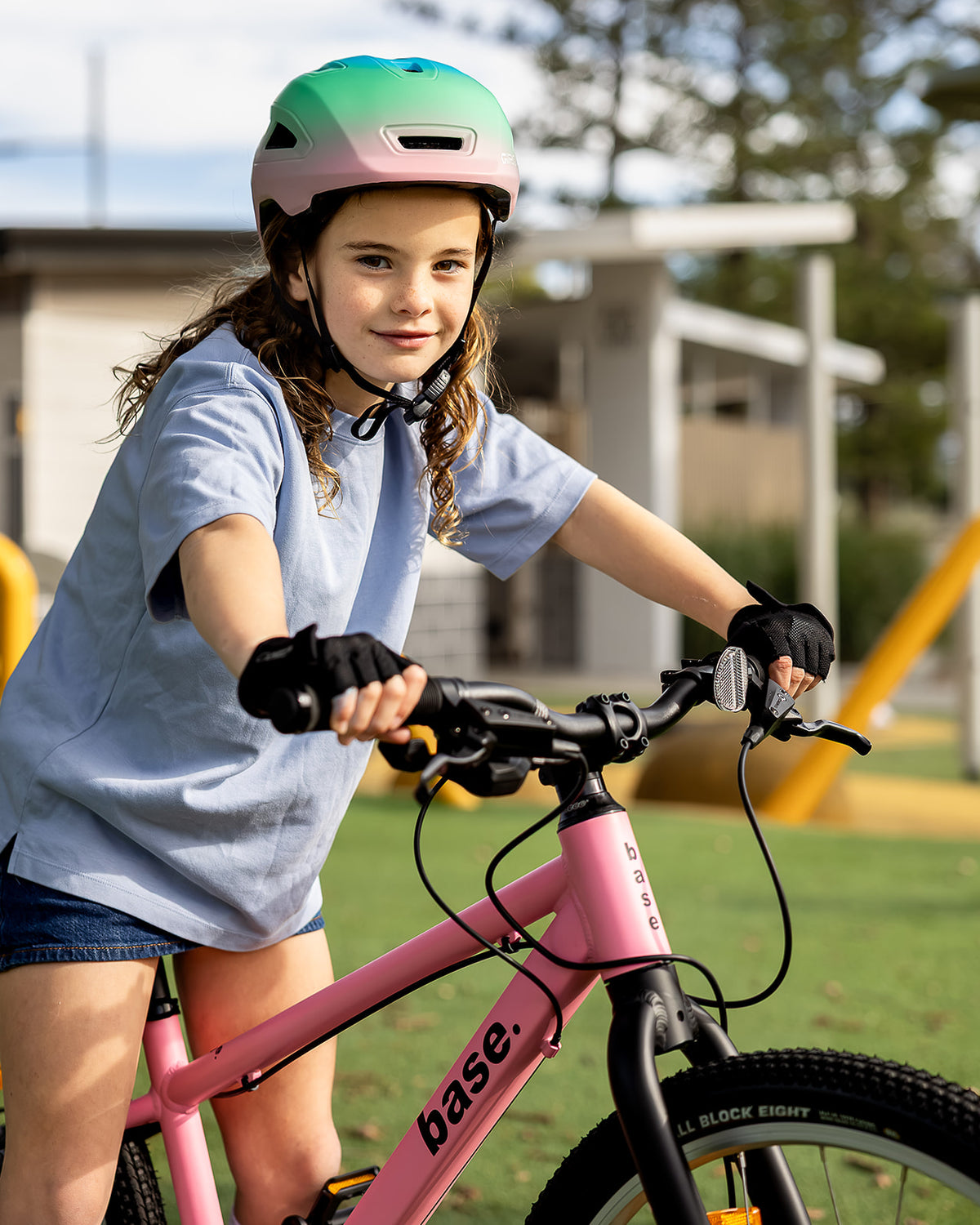 A young girl wearing a pink and green helmet, light blue t-shirt, and black gloves rides a pink base brand bicycle in an outdoor playground setting.
