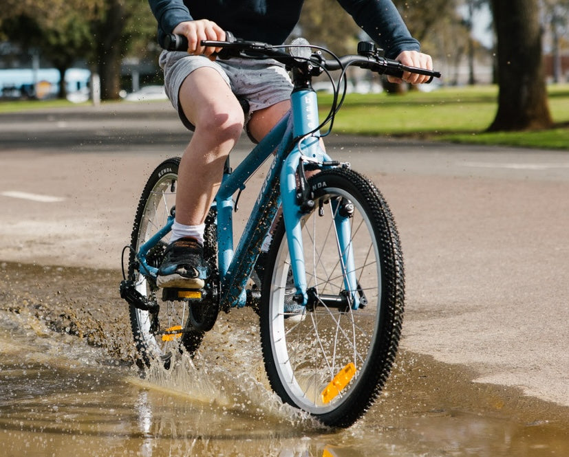 Young boy riding his blue 20 inch push bike through a large puddle creating a splash