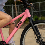 A close-up lifestyle shot of a person riding a pink and black bicycle outdoors. The frame of the bike has the brand name base.