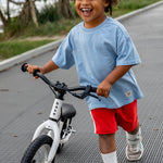 A young child with curly hair riding a white balance bike on a paved path outdoors, wearing a light blue t-shirt, red shorts, white socks, and sneakers.