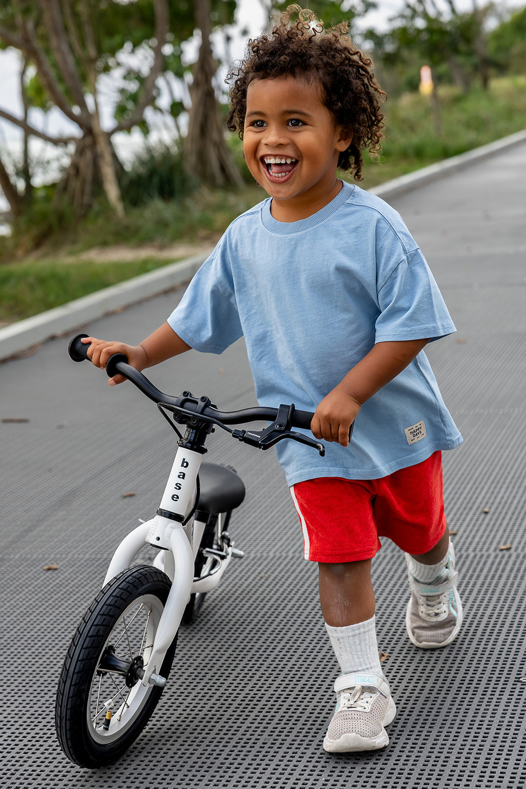 A young child with curly hair riding a white balance bike on a paved path outdoors, wearing a light blue t-shirt, red shorts, white socks, and sneakers.